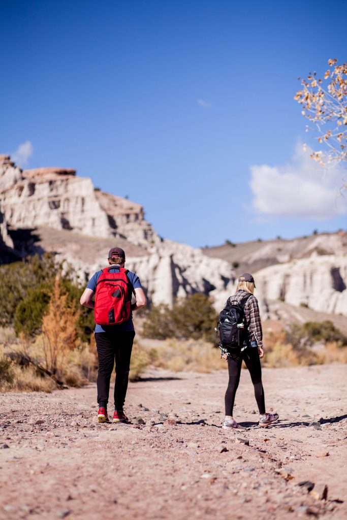 A young couple stands in a mountainous desert with backpacks about to begin their hike. 