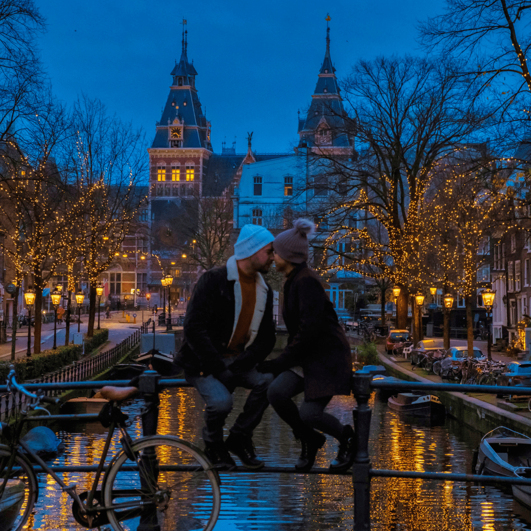 Couple shares a moment on an Amsterdam canal bridge with festive lights and a historic building backdrop at dusk.