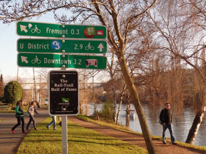 Mileage signs on the Burke Gilman Trail with people walking along the water in the background.