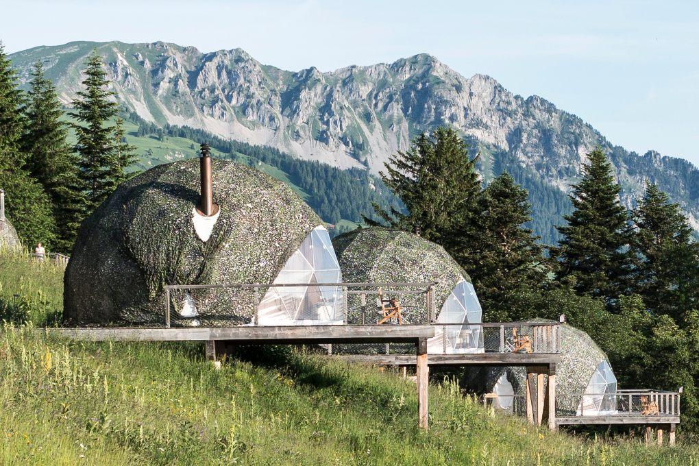 Three geodesic pods line a hillside with Swiss Alps in the background.