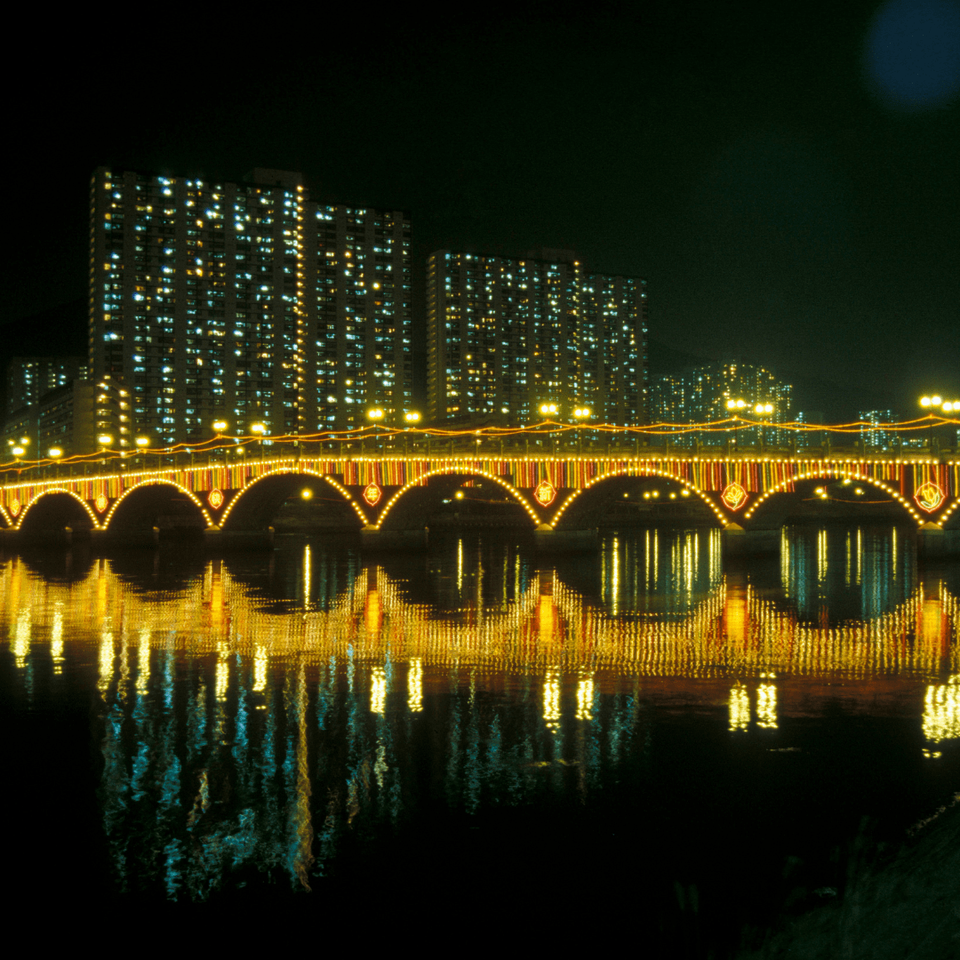 Illuminated bridge and high-rises reflect on water at night in Kowloon, Hong Kong, showcasing urban vibrancy and festive lights.