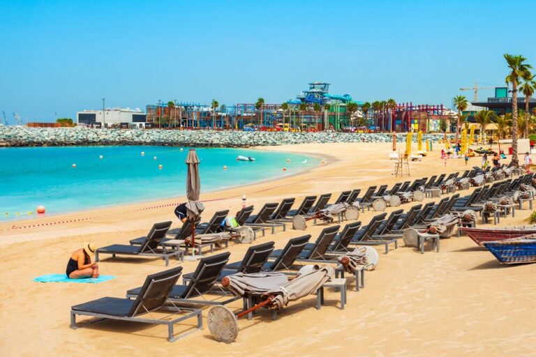 Sun loungers line the sandy shore of La Mer Beach with a waterpark in the distance.