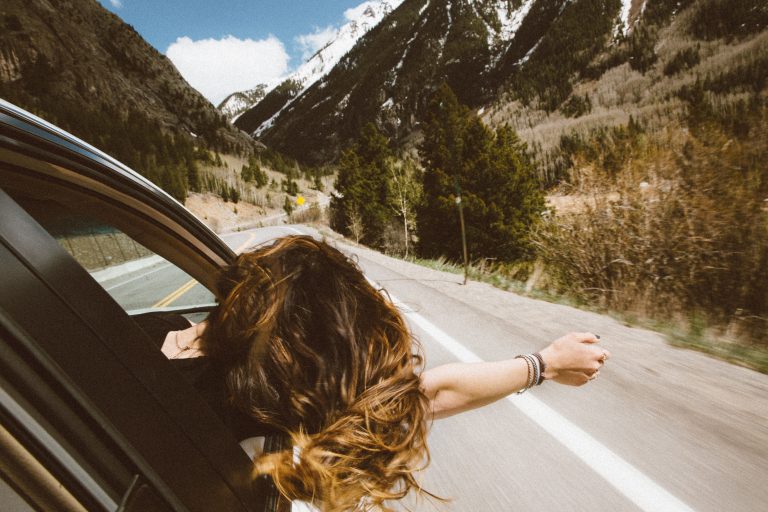 Woman sticking her head out of car window on highway during roadtrip through mountains.